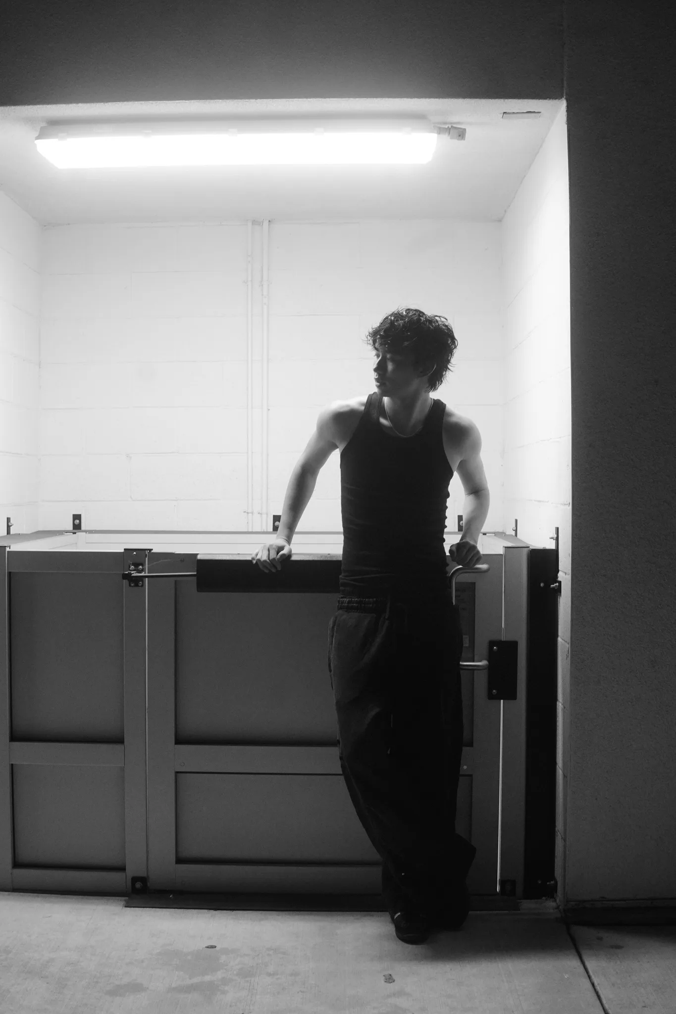 Black-and-white portrait of a model in a black tank leaning on a parking gate beneath a fluorescent light