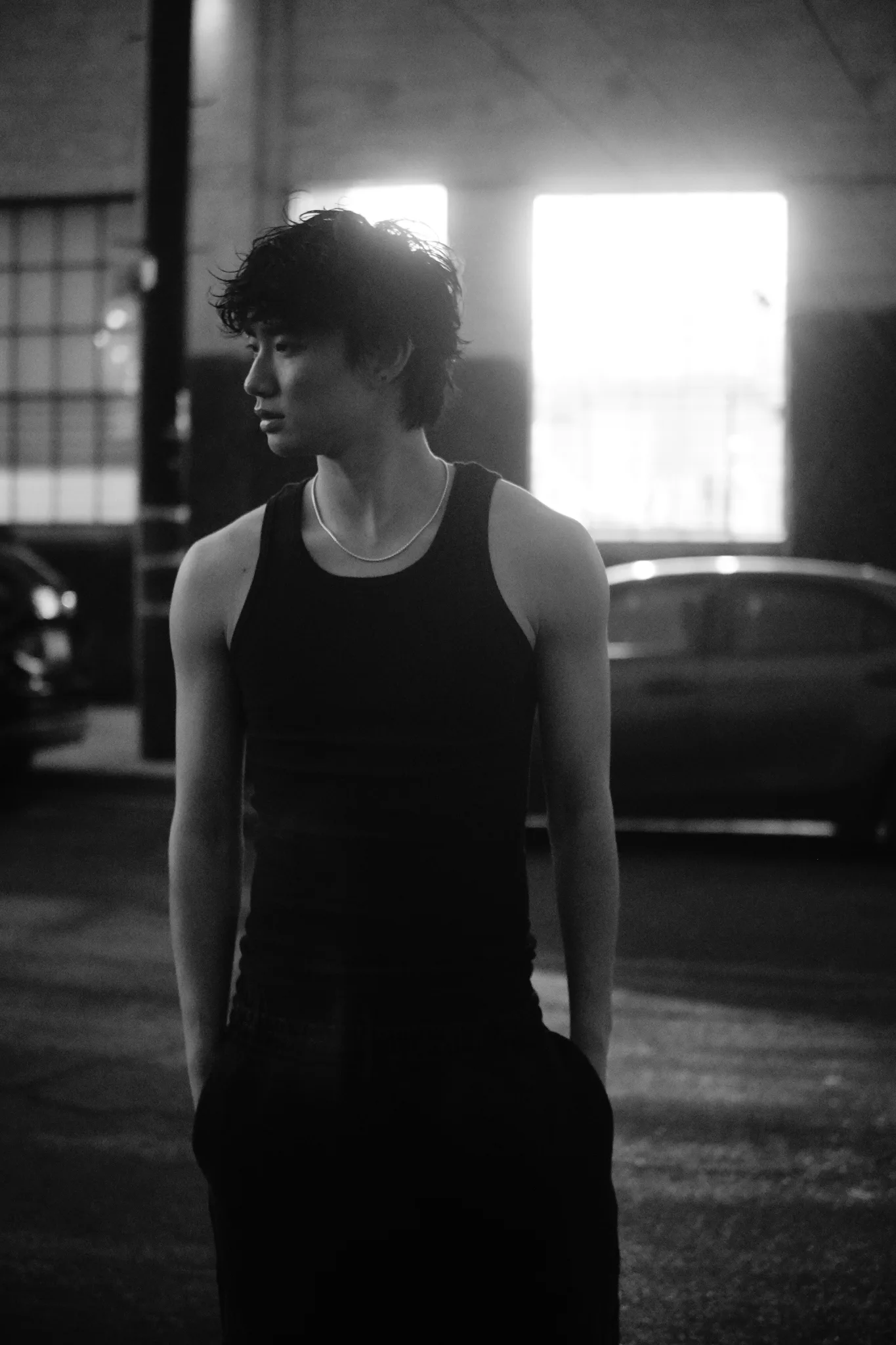Black-and-white backlit portrait of a model in a black tank and chain necklace on a dark warehouse street