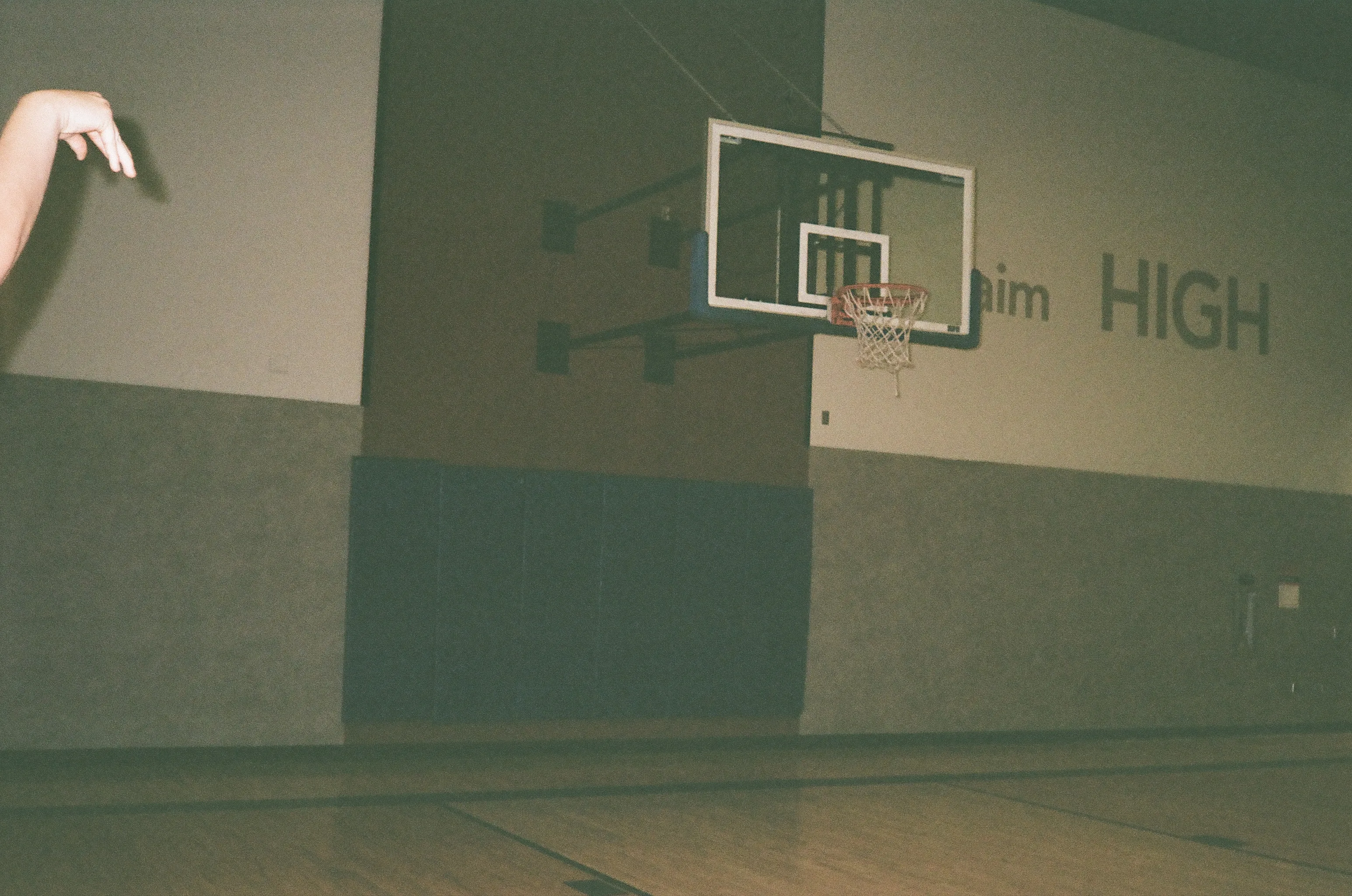 Flash-lit basketball hoop in a school gym beneath painted 'HIGH' lettering, a hand reaching in from the edge of the frame