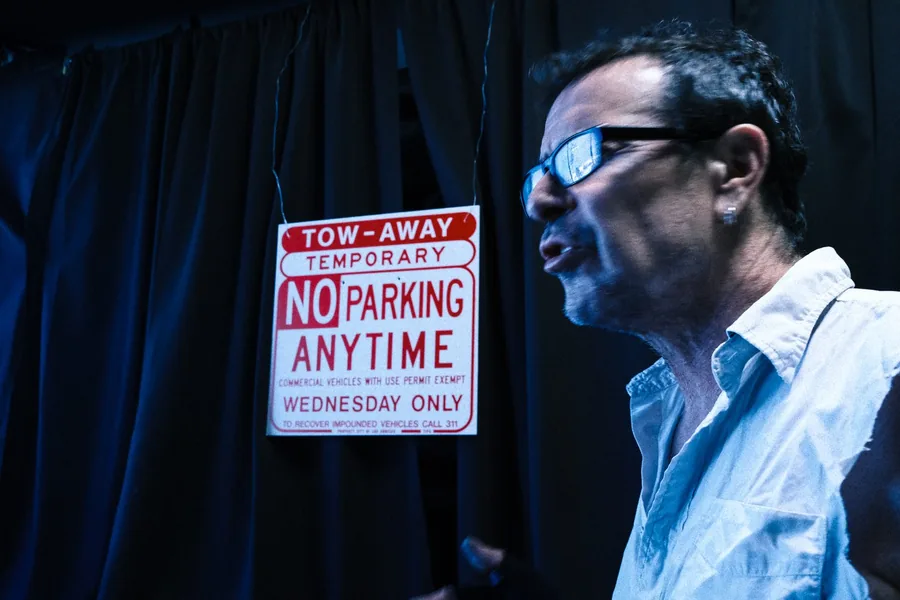Man in glasses beside a temporary 'No Parking Anytime' tow-away sign, lit in cold blue light