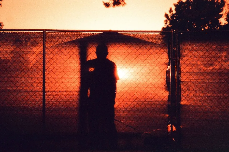 Silhouette standing at a chain-link fence against a deep orange sunset