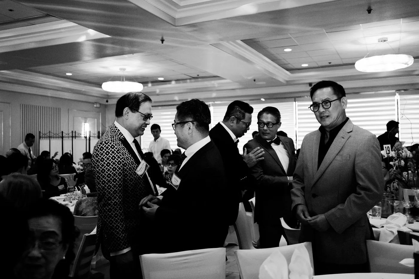 Black-and-white banquet-hall scene of men in suits greeting each other between tables
