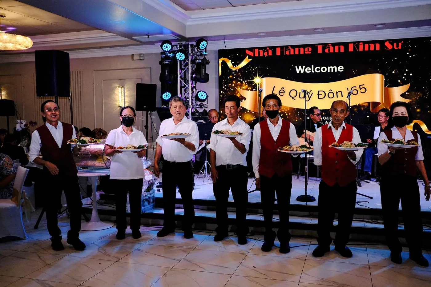 Waiters in vests presenting plates before a 'Welcome Lê Quý Đôn '65' banquet stage
