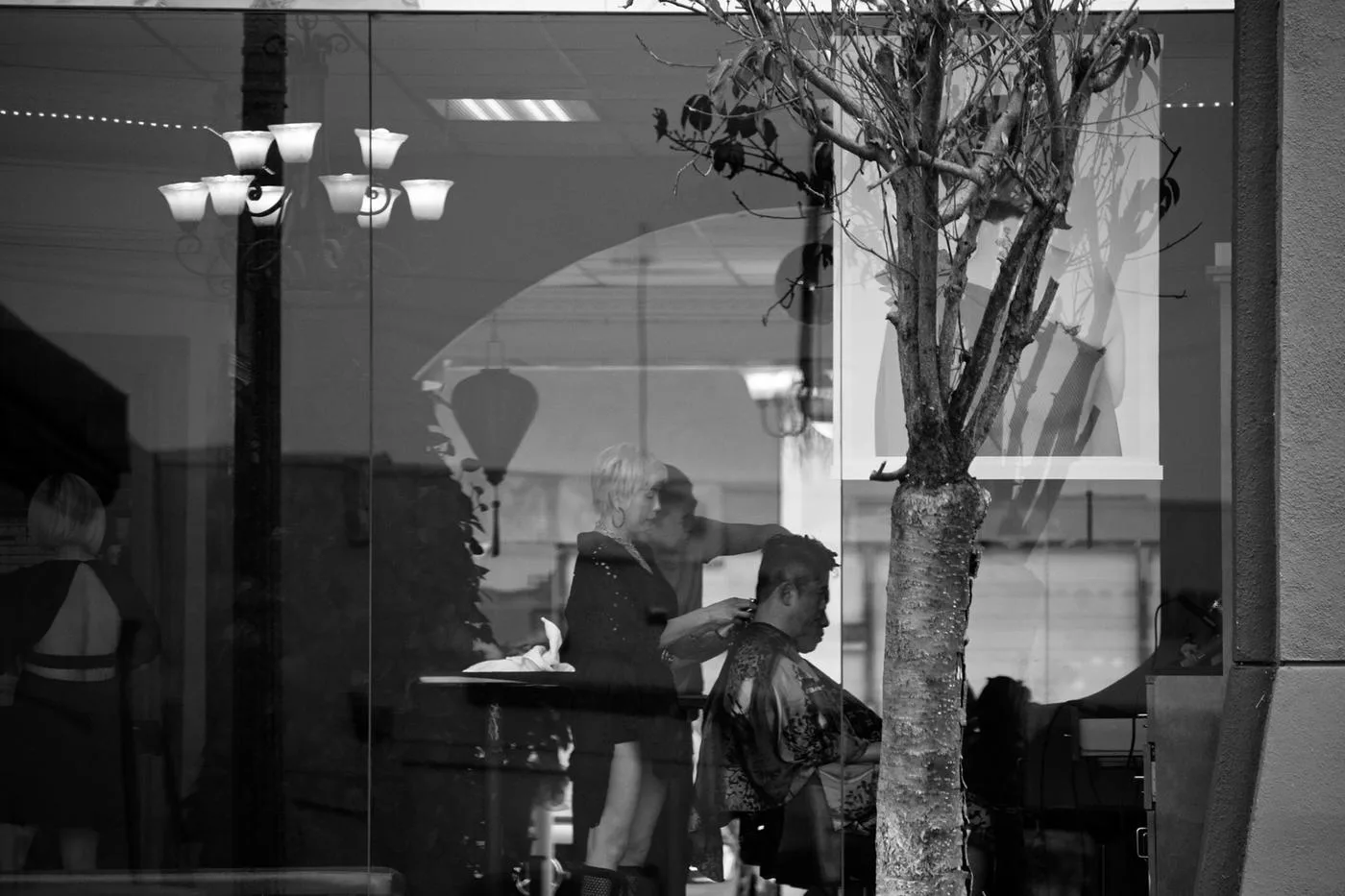 Black-and-white view through a barbershop window of a client in a cape and stylist at work