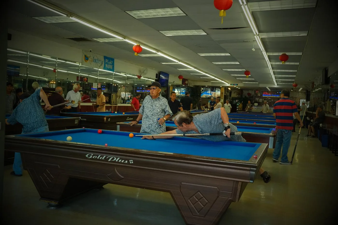 Men playing pool under red lanterns in a crowded billiards hall