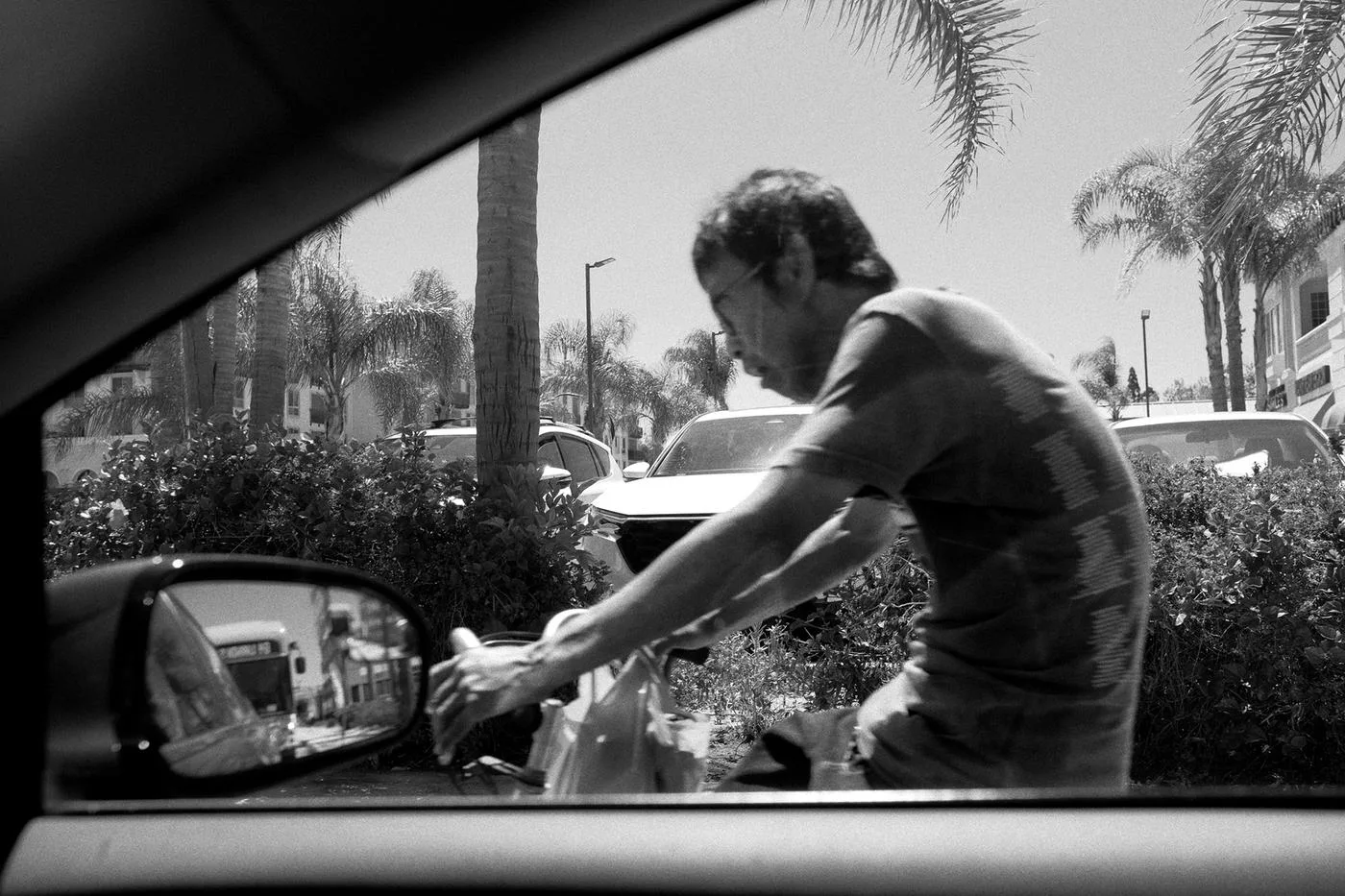 Black-and-white frame through a car window of a man loading groceries between palm trees