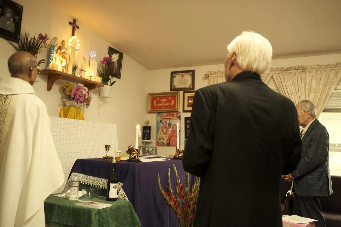 Priest and elders praying at a home altar with a crucifix, candles, and framed icons