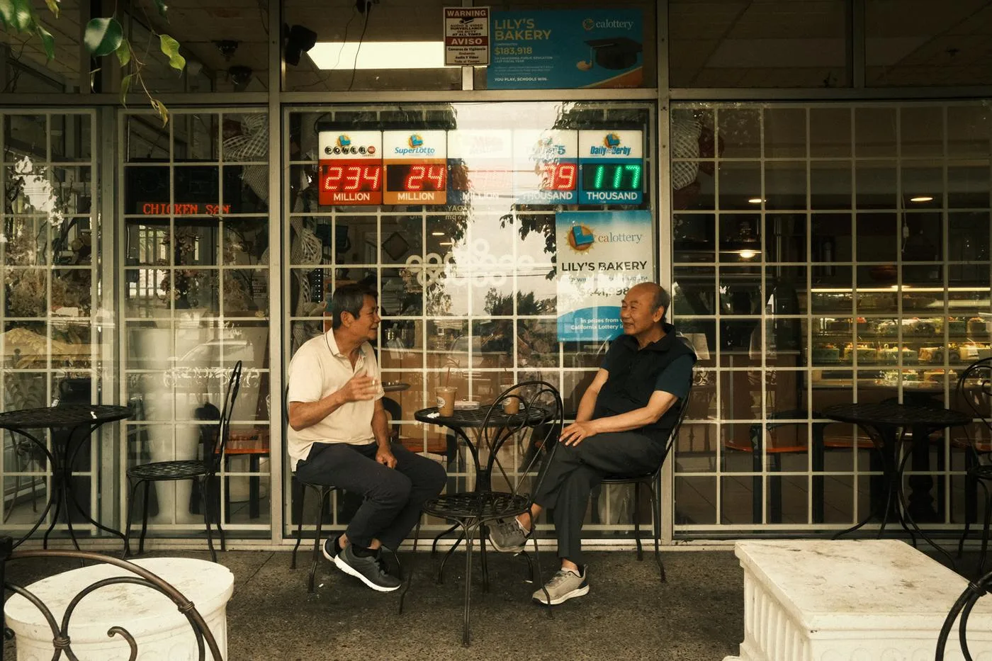 Two men talking at a table outside Lily's Bakery beneath lottery jackpot signs