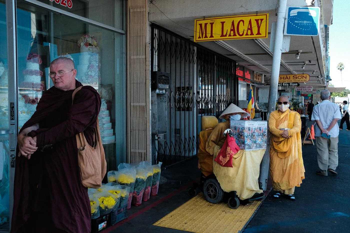 Monk in robes outside Mi La Cay bakery as nuns tend a cart, cut flowers for sale beside