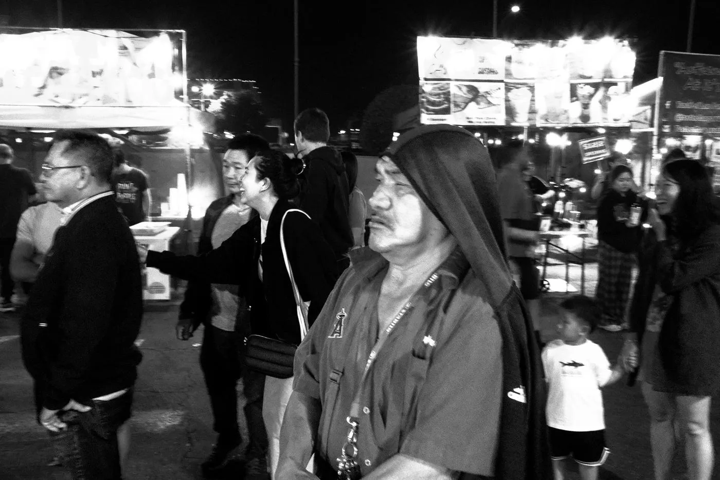 Black-and-white night-market crowd, an older man in a hood passing lit food stalls