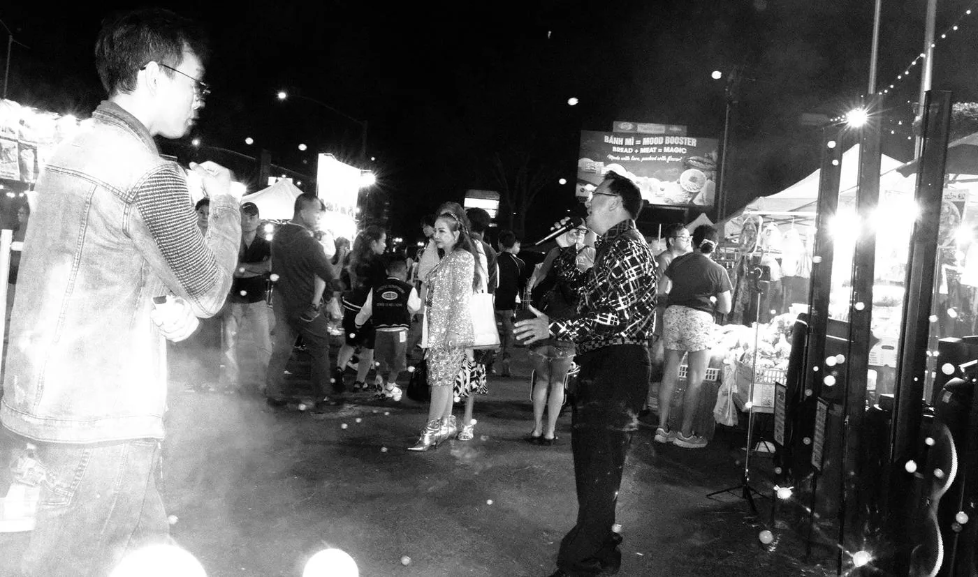 Black-and-white night-market performance, a singer holding the mic mid-song between stalls