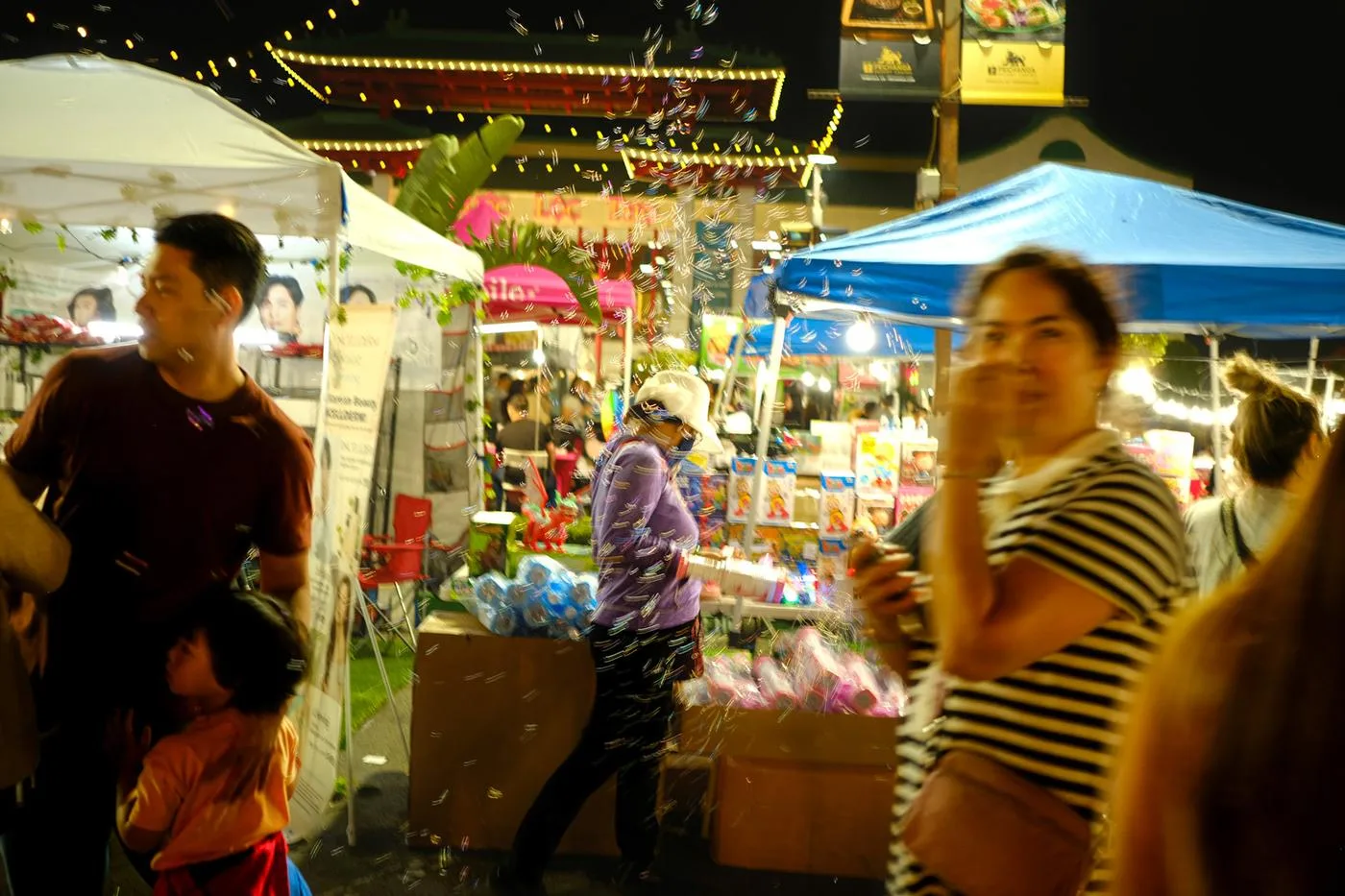 Night-market lane with foam snow drifting over shoppers between stalls