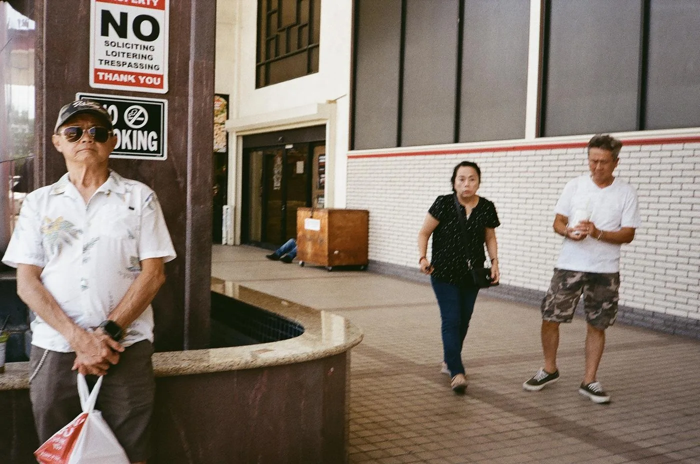 Older man in sunglasses beneath 'No Soliciting' signs as a couple walks past white brick