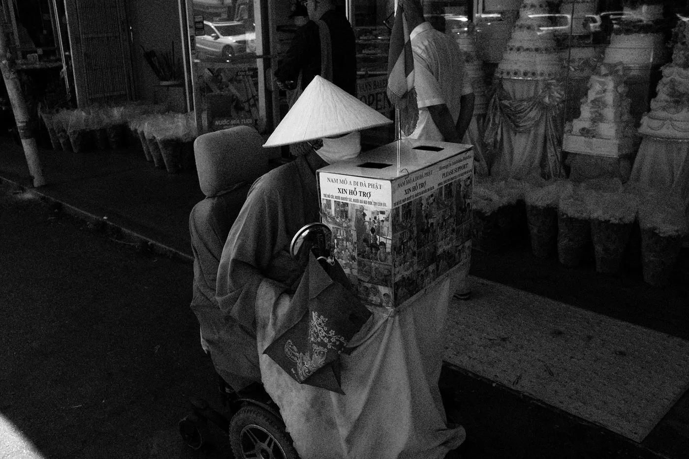 Black-and-white photo of a vendor in a nón lá conical hat on a mobility scooter with a donation box outside a shop