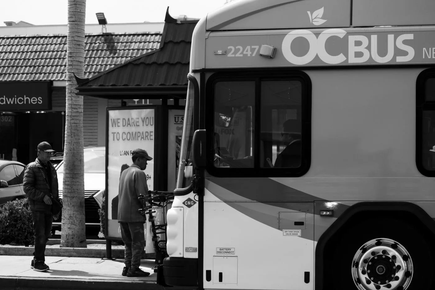Black-and-white frame of an OC Bus at a stop as a rider loads a bicycle onto the front rack