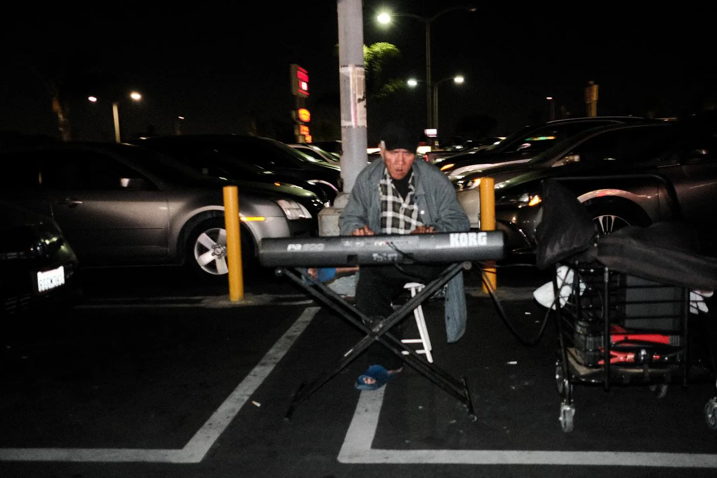 Man in a hat playing a keyboard set up between cars in a parking lot at night