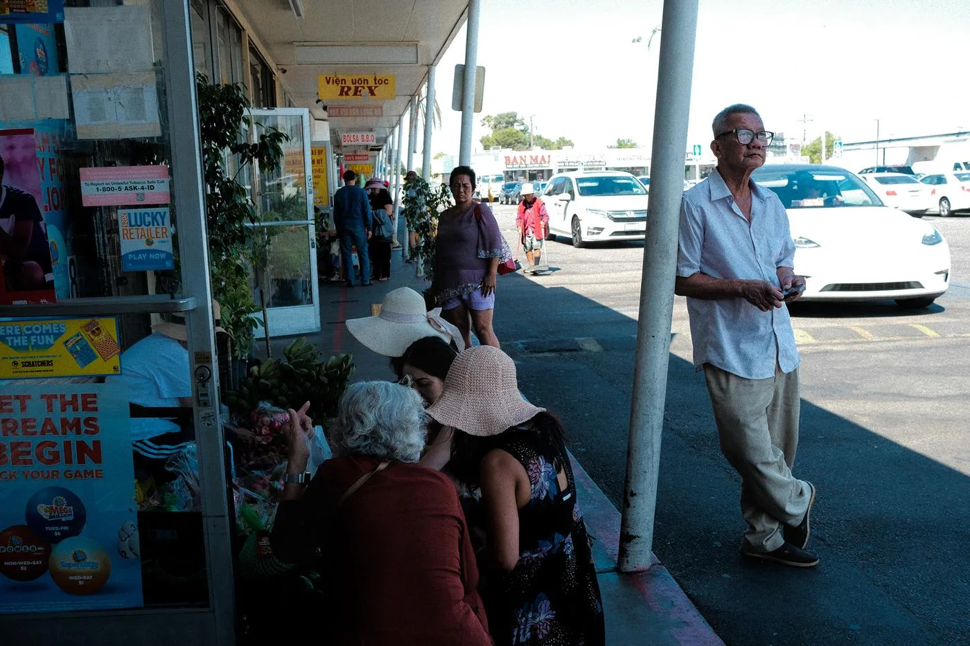 Women in sun hats gathered on a plaza curb outside shops, a man walking past