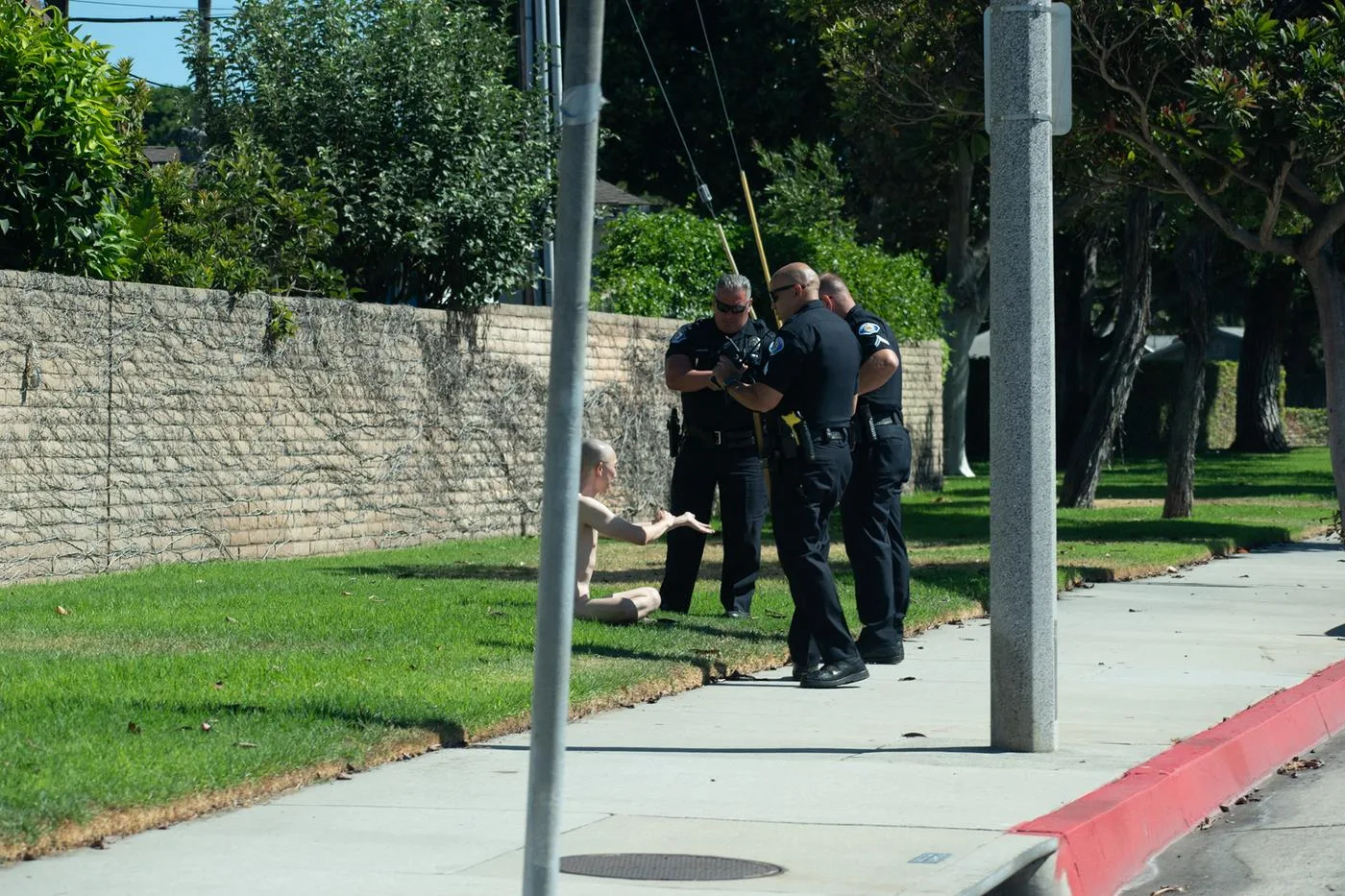 Three police officers speaking with a man seated on a lawn beside the sidewalk