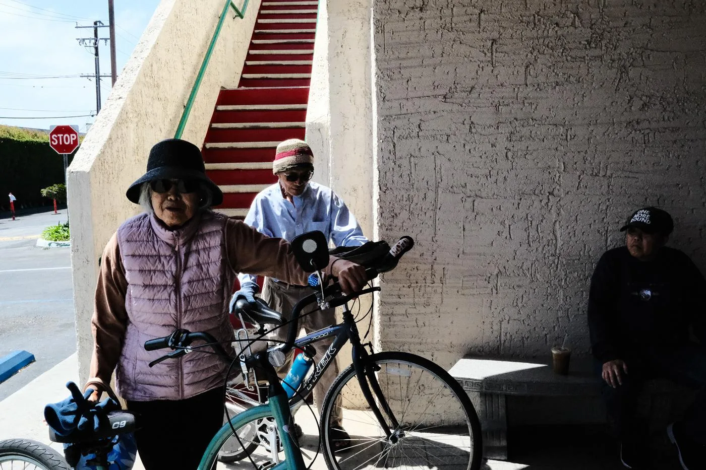 Woman in a bucket hat wheeling a bicycle past red-carpeted stairs, two men resting behind