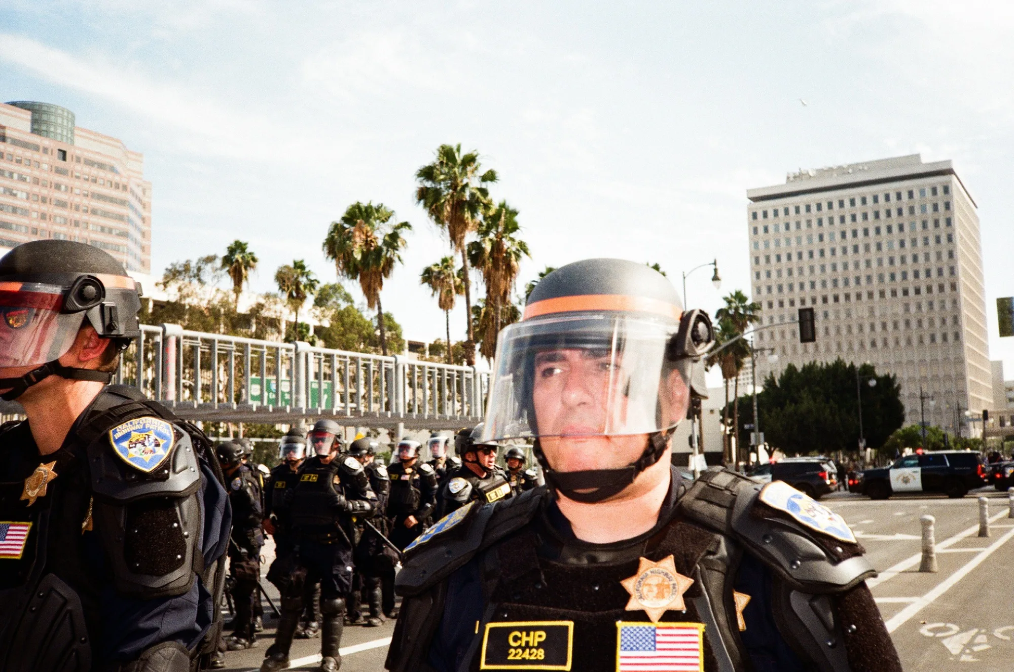 CHP officers in riot helmets lined up before palm trees and civic buildings