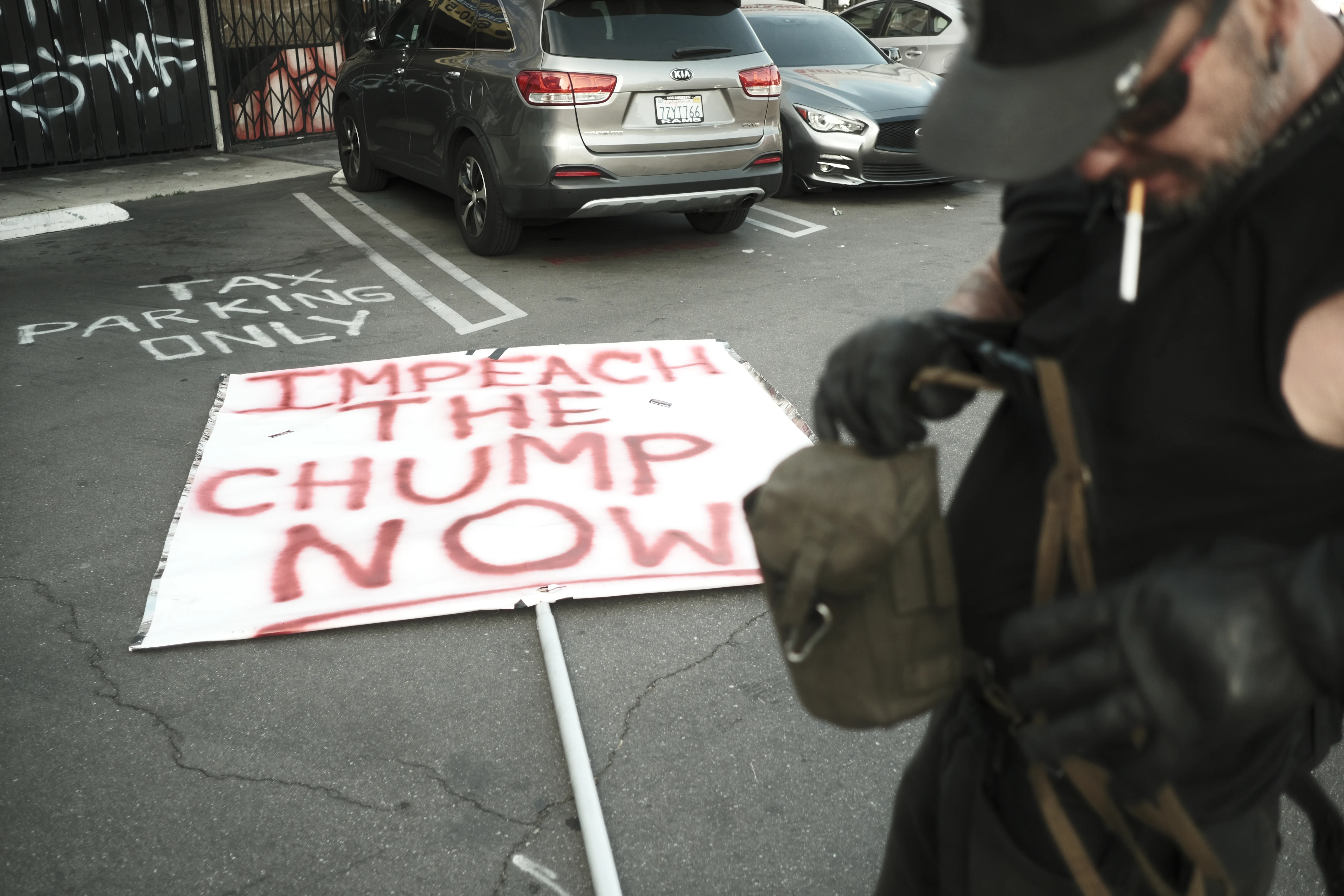 Protester in black with a cigarette beside a hand-painted 'Impeach the Chump Now' sign lying in a parking lot