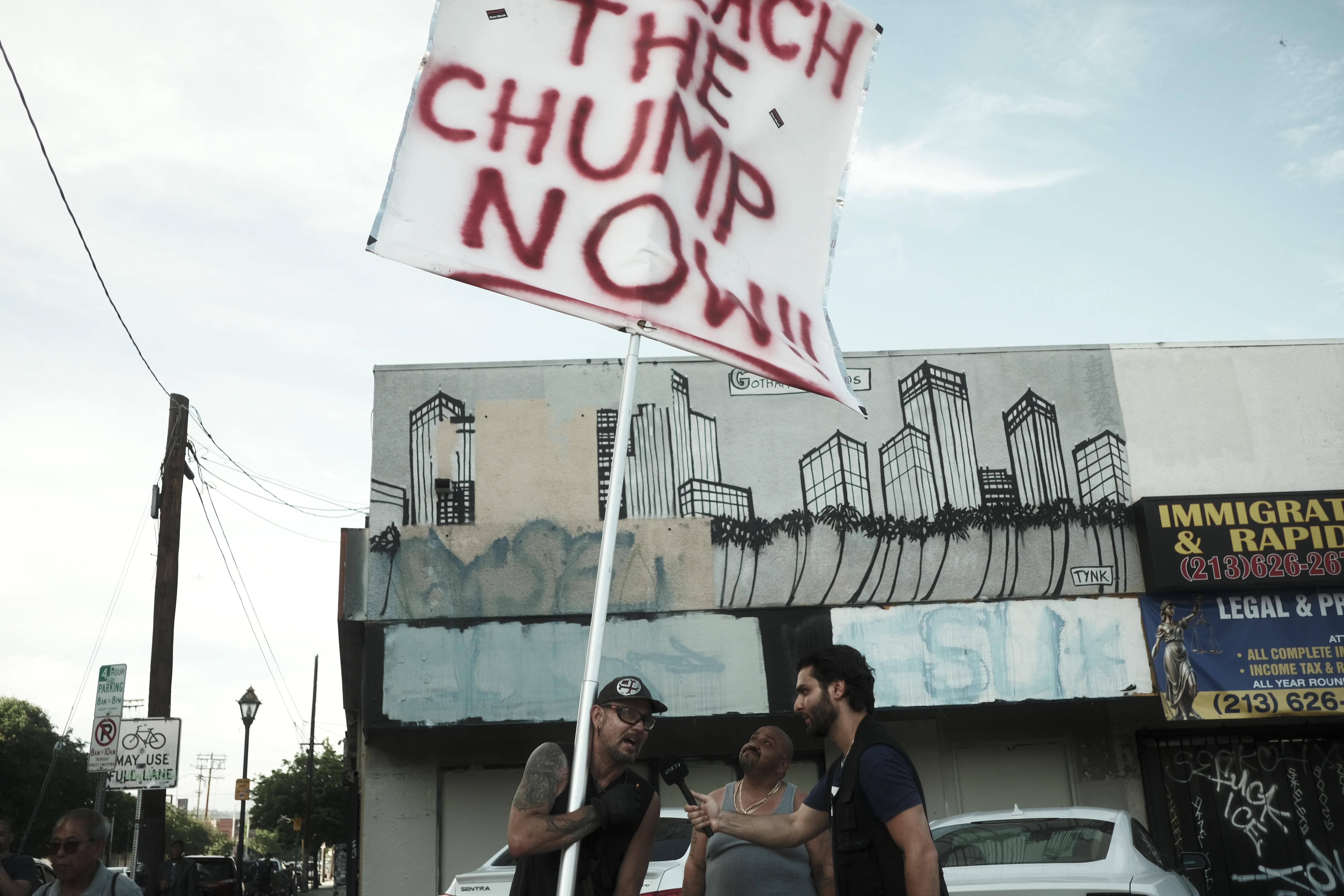Protesters talking beneath a raised 'Impeach the Chump Now' sign on a muraled street corner