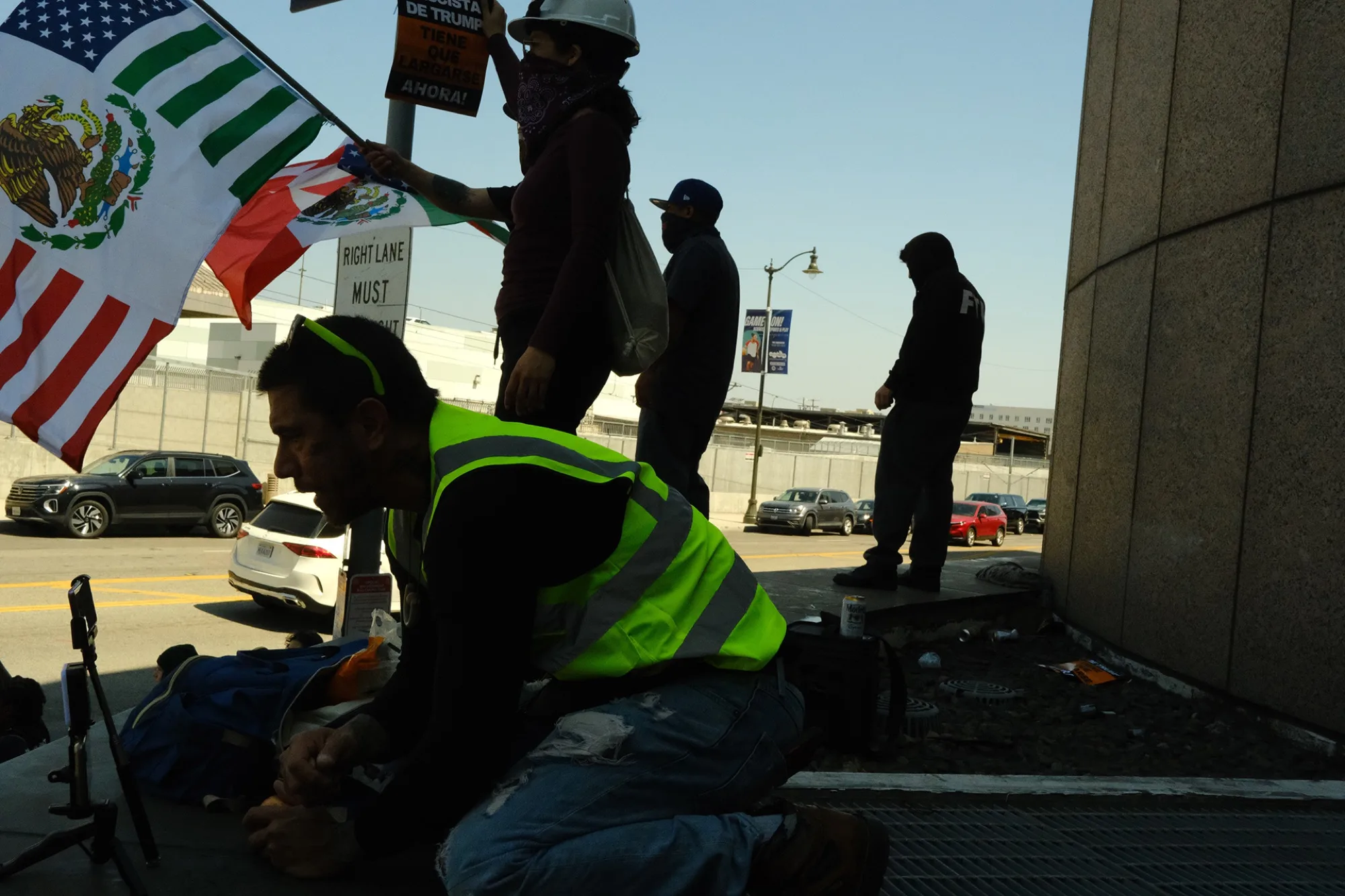 Protester in a safety vest kneeling beside Mexican and American flags outside a transit center